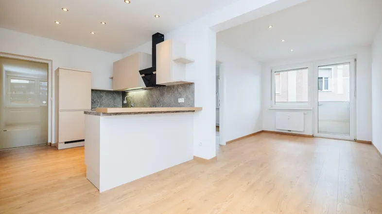 Heller offener Wohnbereich mit moderner Einbauküche, Holzboden und Zugang zum Balkon.
