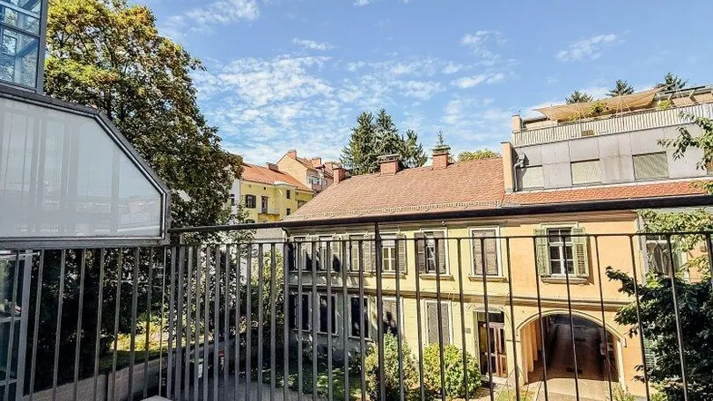 Balkon mit Blick auf einen begrünten Innenhof und umliegende Gebäude unter blauem Himmel.