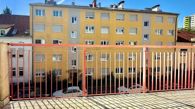 Heller Balkon mit rotem Geländer und Blick auf ein gegenüberliegendes Wohngebäude unter blauem Himmel.