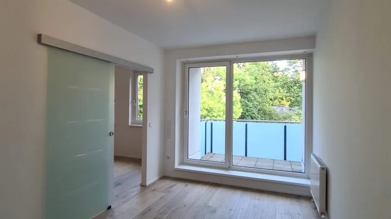 Helles Zimmer mit Holzboden, Schiebetür und Zugang zu einem Balkon mit Blick ins Grüne.