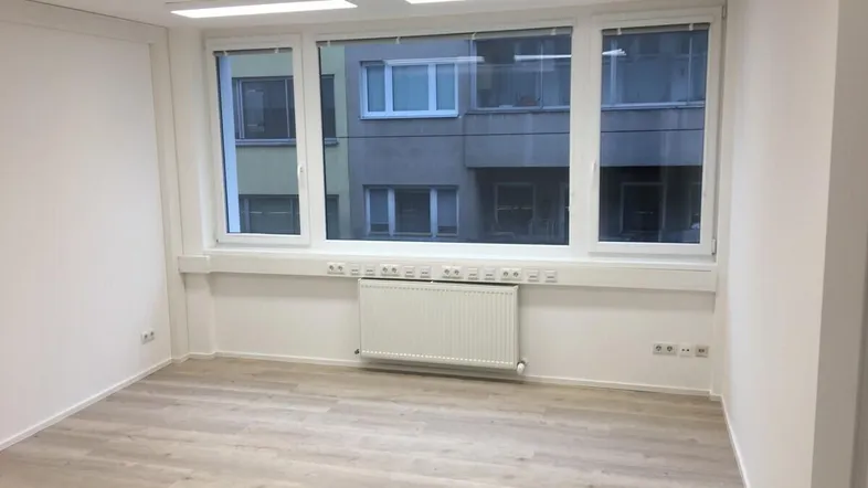 Heller Büroraum mit großen Fenstern, hellem Laminatboden und Heizkörper unter der Fensterbank.