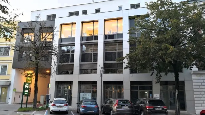 Modernes Geschäftsgebäude mit großen Fenstern und Parkplätzen davor, beleuchtet am Abend.