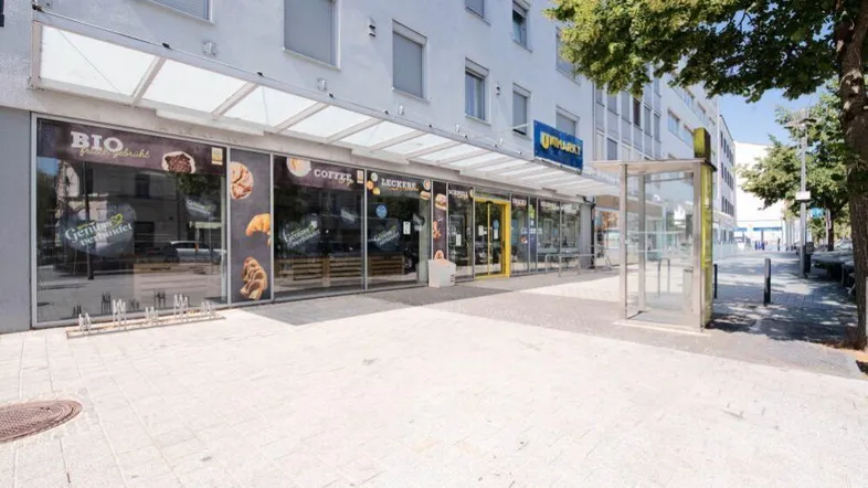 Moderne Ladenfront eines Supermarktes mit großen Glasfenstern und einem Vordach, direkt an einem Gehweg gelegen.