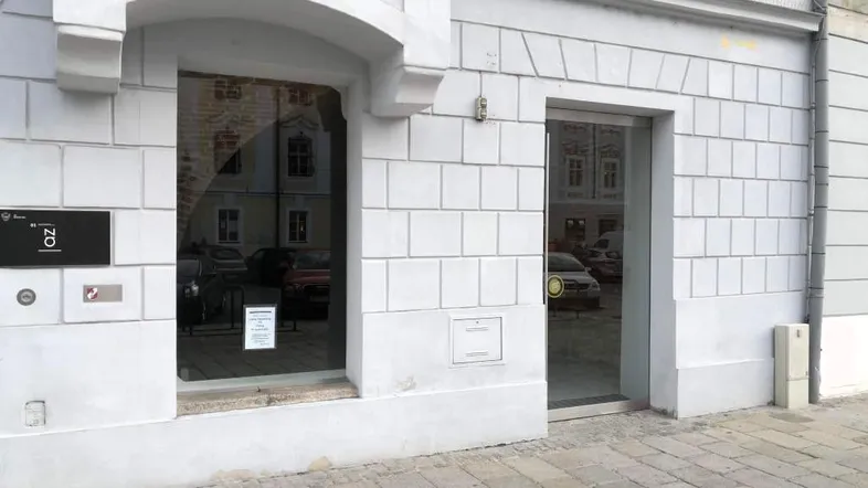 Helle Fassade eines Gebäudes mit großem Schaufenster und gläserner Eingangstür, die Reflexionen der Umgebung zeigt.