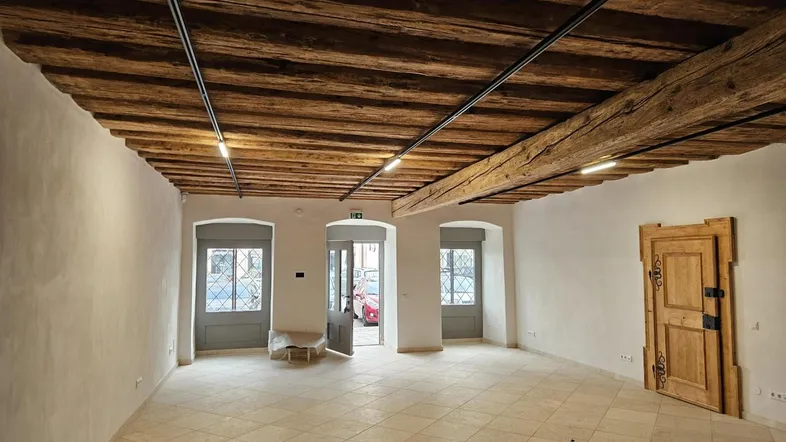 Geräumiger, leerer Raum mit beeindruckender Holzbalkendecke, hellen Fliesen und mehreren Fenstern zur Straße.