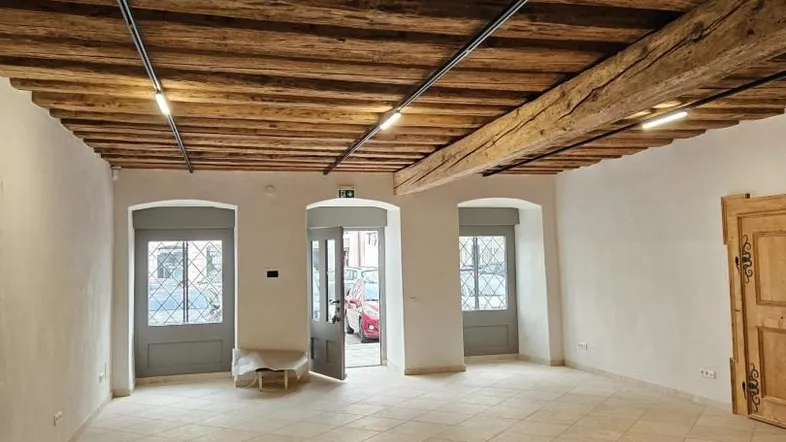Geräumiger Innenraum mit freiliegenden Holzbalken an der Decke und hellen Fliesenböden, gut beleuchtet durch Tageslicht.