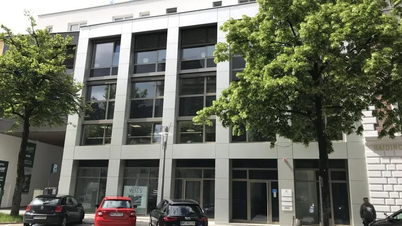 Modernes Geschäftsgebäude mit großen Fenstern und geparkten Autos auf dem Vorplatz unter blauem Himmel.