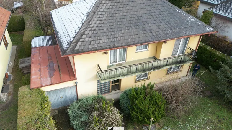 Luftaufnahme eines Einfamilienhauses mit gelber Fassade, grauem Dach und kleinem Balkon, umgeben von Bäumen.