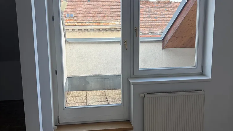 Helles Zimmer mit Parkettboden, Heizkörper und Zugang zu einem kleinen Außenbereich mit Dachblick.