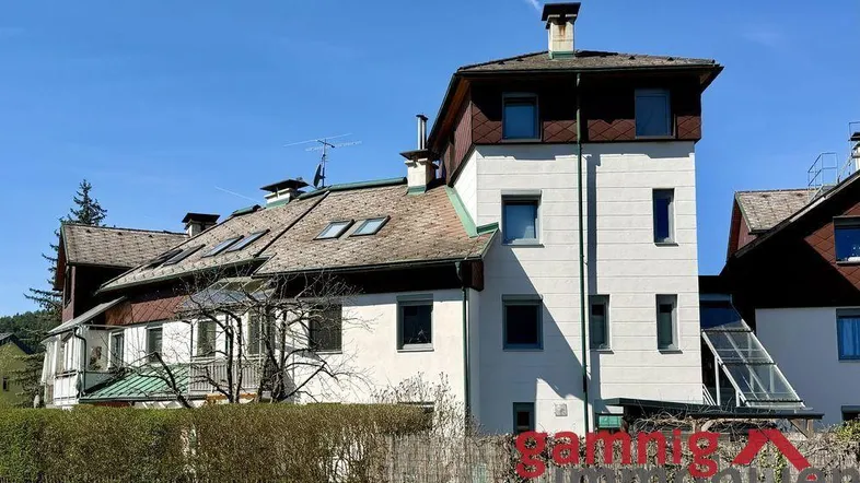 Mehrstöckiges Gebäude mit weißer Fassade, braunen Dachelementen und umgebender grüner Bepflanzung.