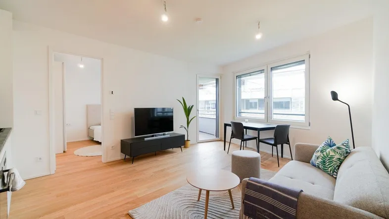Helles Wohn- und Esszimmer mit Holzboden, Zugang zum Balkon und Blick in ein Schlafzimmer.