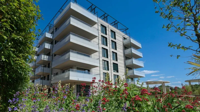 Moderne Wohnanlage mit mehreren Balkonen, umgeben von blühenden Blumenbeeten unter blauem Himmel.