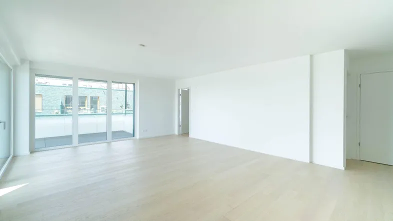 Helles, leeres Wohnzimmer mit großen Fenstern, hellem Parkettboden und Zugang zum Balkon.