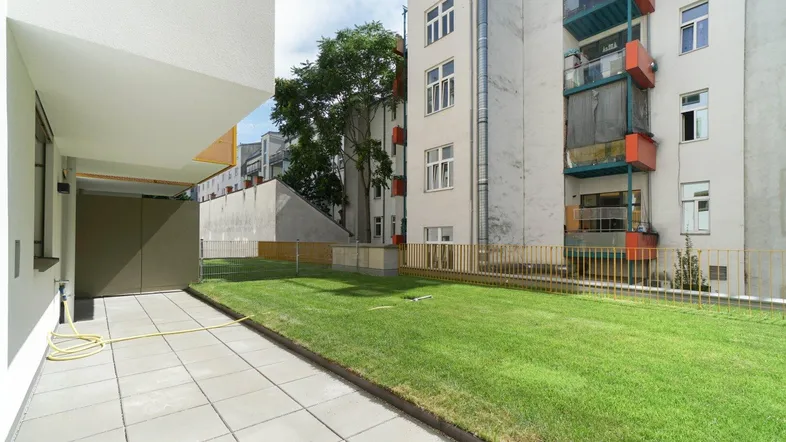 Gepflasterte Terrasse neben einer Rasenfläche mit Blick auf umliegende Gebäude und viel Grün.