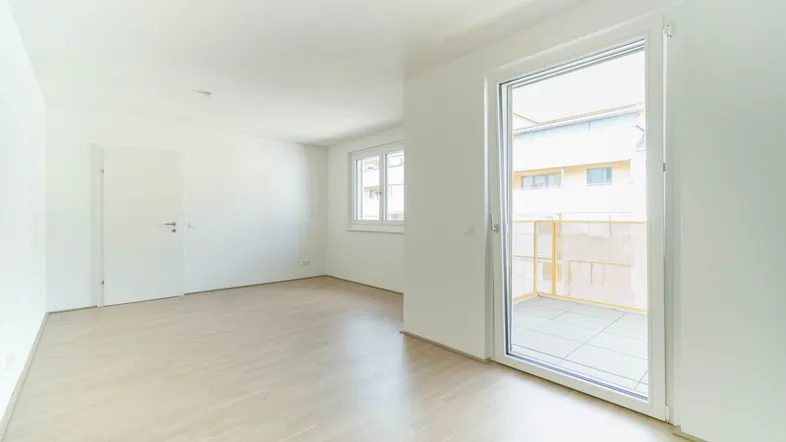 Helles, leeres Zimmer mit hellem Parkettboden und Zugang zu einem Balkon.