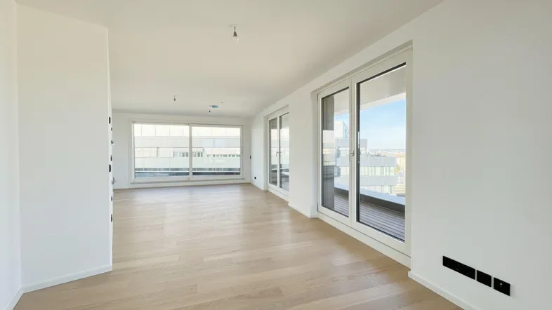 Helles, leeres Wohnzimmer mit Holzboden und großen Fenstern, die viel Tageslicht hereinlassen.