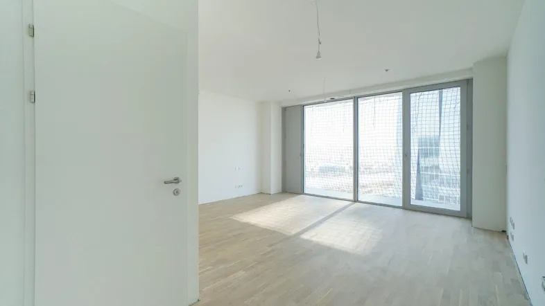 Helles, leeres Zimmer mit Parkettboden und großen Schiebetüren, die viel Tageslicht hereinlassen.