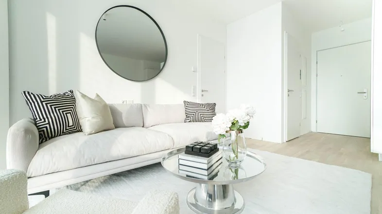 Helles Wohnzimmer mit modernem Sofa, Couchtisch und rundem Spiegel an der weißen Wand.