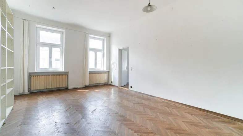 Helles, leeres Zimmer mit Fischgrätparkett und zwei großen Fenstern, bereit zur individuellen Gestaltung.