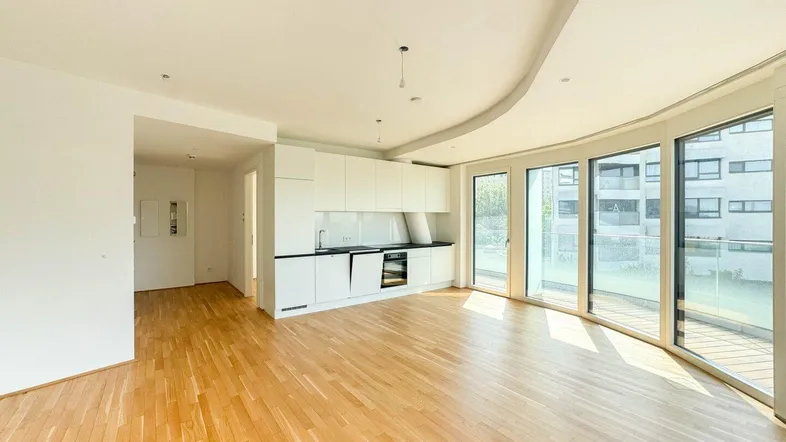 Heller, offener Wohnbereich mit integrierter Küche, Parkettboden und großen Fenstern zum Balkon.