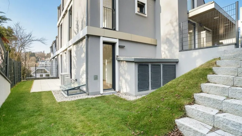 Moderne Gebäudeseite mit kleiner Rasenfläche, Steintreppe und gepflasterter Terrasse.