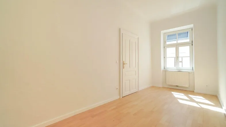 Helles Zimmer mit Parkettboden und einem Fenster, das viel Tageslicht hereinlässt.