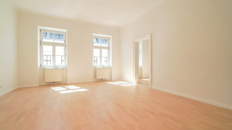 Helles, leeres Zimmer mit Parkettboden und zwei Fenstern, das viel Tageslicht hereinlässt.