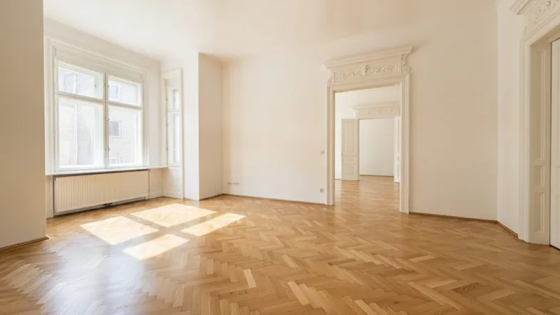 Heller Raum mit Fischgrätparkett, hohen Decken und Stuckverzierungen an der Türöffnung.