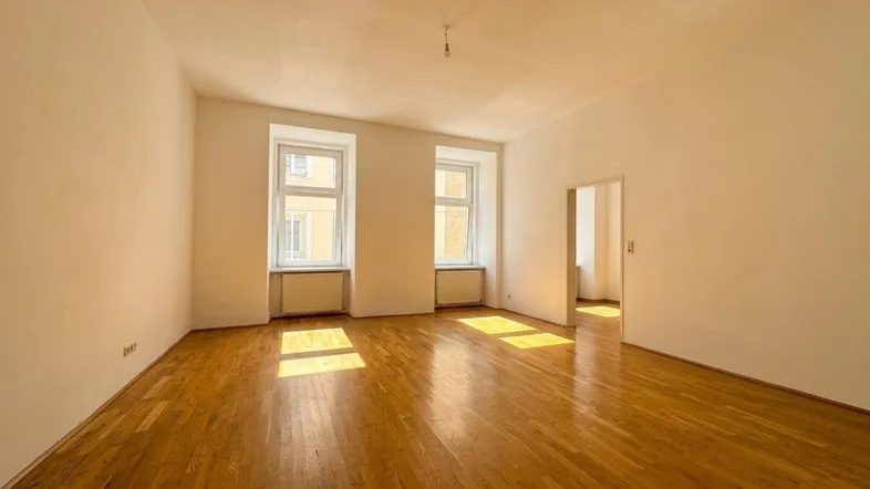 Helles, leeres Zimmer mit Holzboden und zwei großen Fenstern, die viel Sonnenlicht hereinlassen.