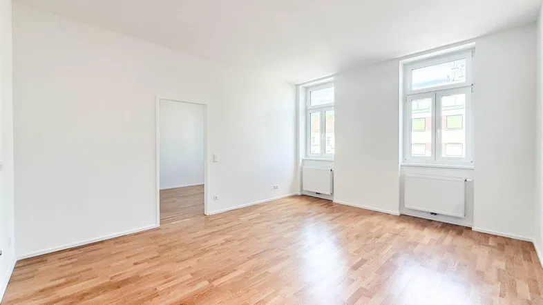 Helles, leeres Zimmer mit Parkettboden und zwei großen Fenstern, bereit zur individuellen Gestaltung.