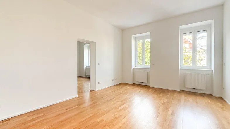 Helles, leeres Zimmer mit Parkettboden und zwei Fenstern, bereit zur individuellen Gestaltung.
