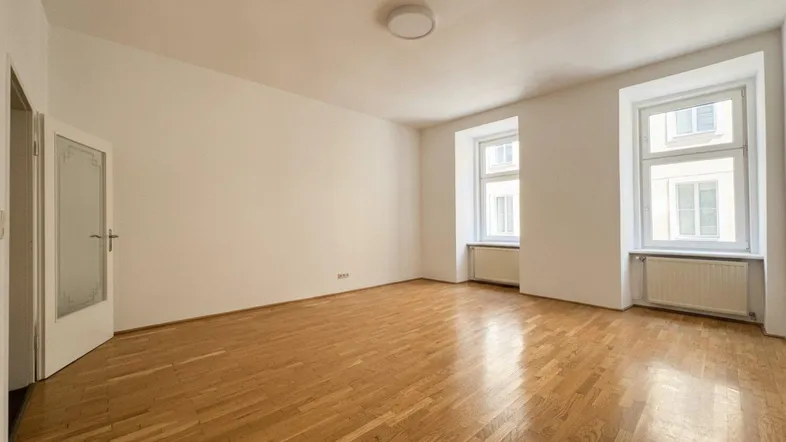 Helles, leeres Zimmer mit Parkettboden und zwei Fenstern, bereit zur individuellen Gestaltung.