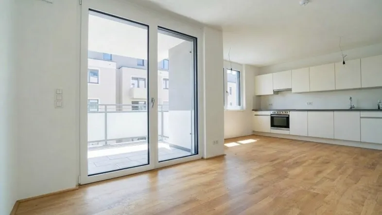 Heller offener Wohnbereich mit integrierter Küche, Holzboden und Zugang zu einem Balkon.