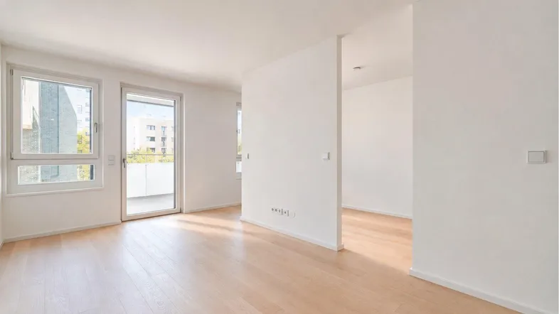 Helles, leeres Zimmer mit Parkettboden, großen Fenstern und Zugang zu einem Balkon.