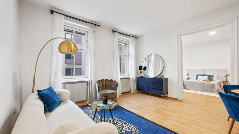 Helles Wohnzimmer mit modernem Sofa, blauer Teppich und Blick in ein Schlafzimmer.