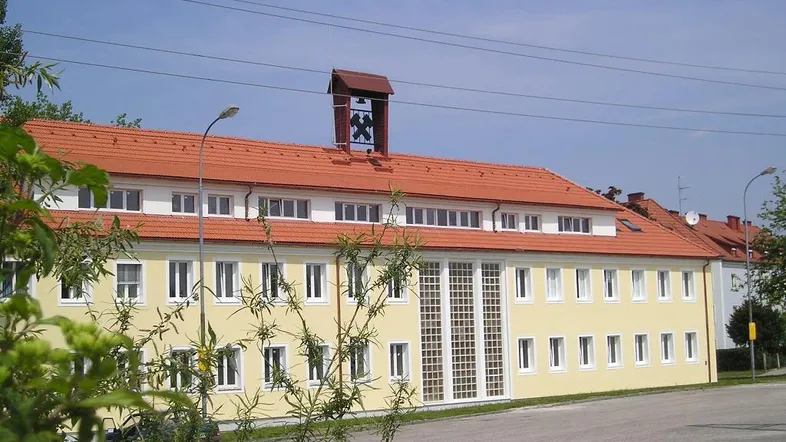 Gebäudeansicht mit gelber Fassade, rotem Ziegeldach und einem Glockenturm unter blauem Himmel.