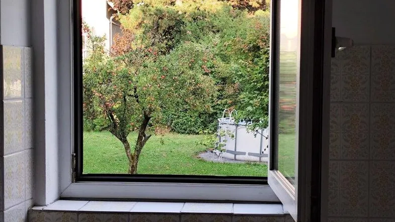 Badezimmer mit geöffnetem Fenster und Blick ins Grüne, umgeben von hellen Fliesen.