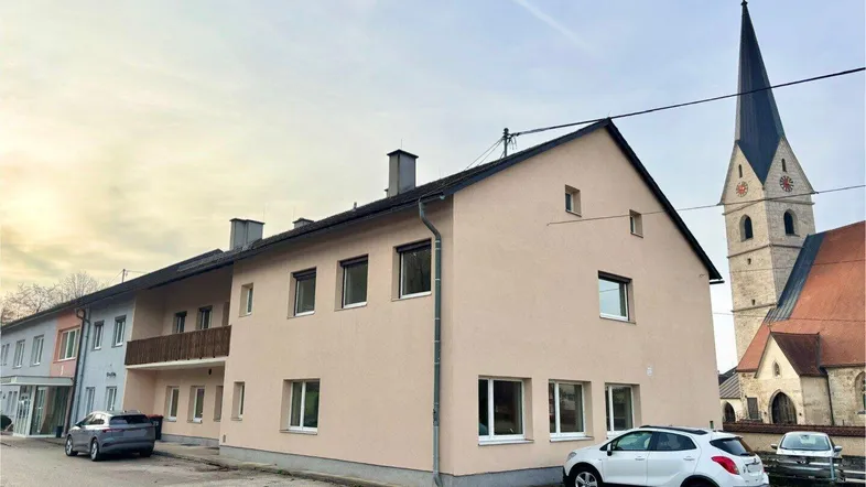 Gebäudeansicht mit beiger Fassade, Satteldach und angrenzender Kirche im Hintergrund.