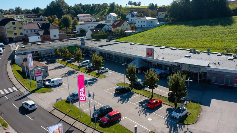 Drohnenaufnahme eines Einkaufszentrums mit großem Parkplatz, umgeben von Wohnhäusern und grünen Feldern.