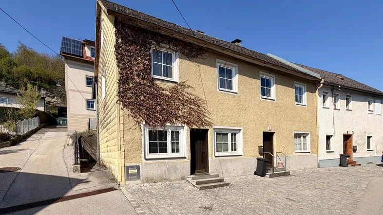 Gelbes Reihenhaus mit bewachsener Fassade und gepflastertem Vorplatz unter blauem Himmel.