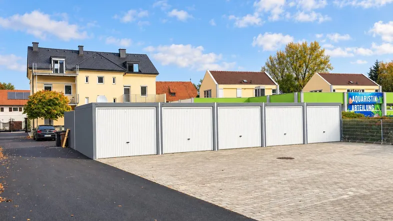 Reihe von Garagenboxen mit weißen Toren und einem gepflasterten Vorplatz unter blauem Himmel.