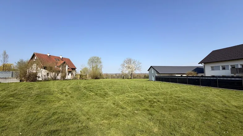 Weitläufige grüne Wiese mit traditionellen und modernen Häusern im Hintergrund und Fernsicht.