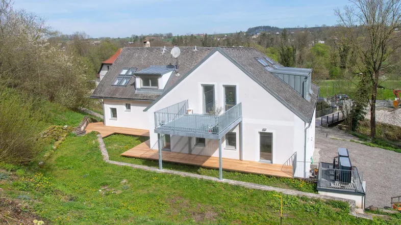 Modernes Haus mit Balkon und Holzterrasse in einer grünen, hügeligen Landschaft.
