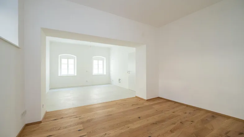 Heller Wohnbereich mit Holzboden und offenem Übergang zu einem weiteren Raum mit Fliesenboden und Fenstern.