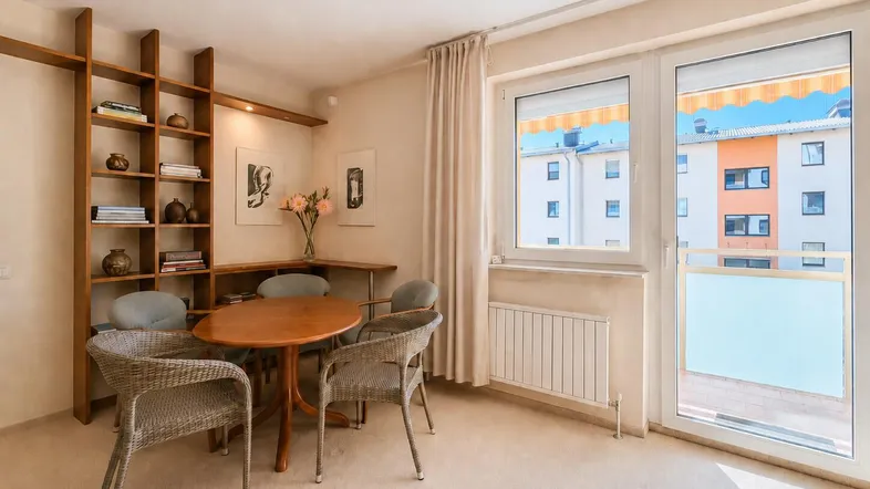 Heller Wohnbereich mit Esstisch, Bücherregal und Zugang zu einem Balkon mit Blick auf umliegende Gebäude.