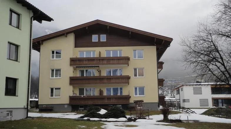 Mehrfamilienhaus mit Balkonen und Satteldach in verschneiter Berglandschaft, umgeben von Grünflächen.