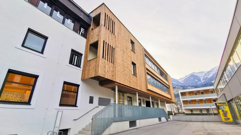 Modernes Gebäude mit weißer Fassade und Holzverkleidung, umgeben von Bergen und einem Parkplatz.