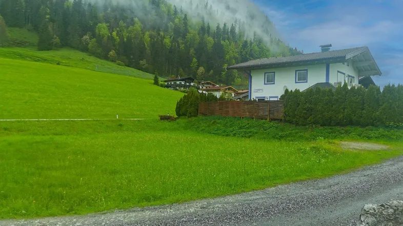Grüne Wiese vor einem Haus mit bewaldetem Hang und nebligen Bergen im Hintergrund.