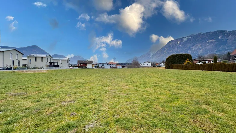 Offene grüne Fläche mit Blick auf eine Siedlung und die umliegenden Berge unter einem weiten Himmel.