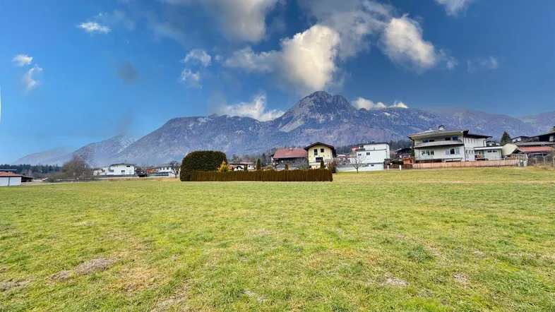 Weitläufige grüne Wiese mit Häusern und Bergen im Hintergrund unter blauem Himmel.